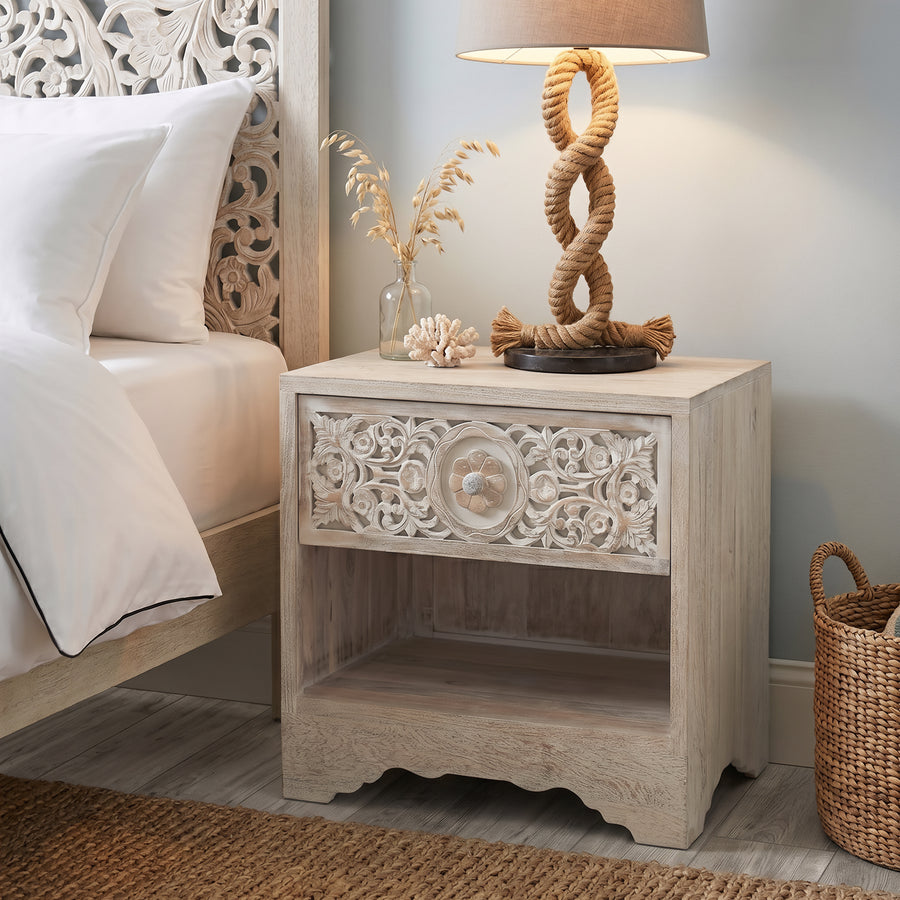 A light beige carved wooden nightstand in a bedroom setting, styled with a rope lamp, dried grass in a glass vase, and a small seashell.
