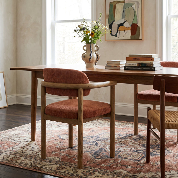 A dining room scene with a wooden table and upholstered chairs. The chairs have warm rust-orange velvet cushions and light natural wood frames.