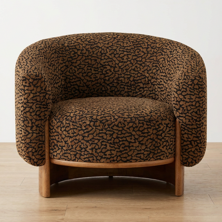 A modern barrel-style armchair with a curved back and arms, upholstered in a brown and black abstract organic patterned fabric, supported by a light natural wood frame and legs, photographed against a neutral white wall and light wood floor.