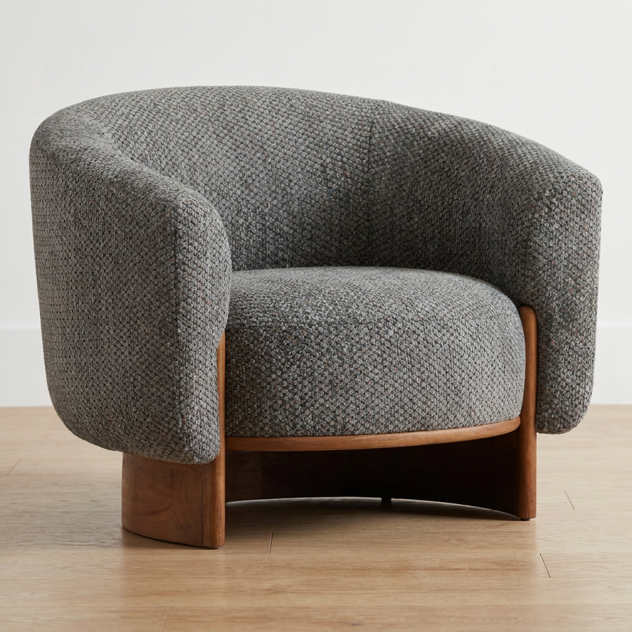 A modern barrel-style armchair upholstered in textured gray woven fabric with a curved wooden base, shown on a light wood floor against a plain white wall.