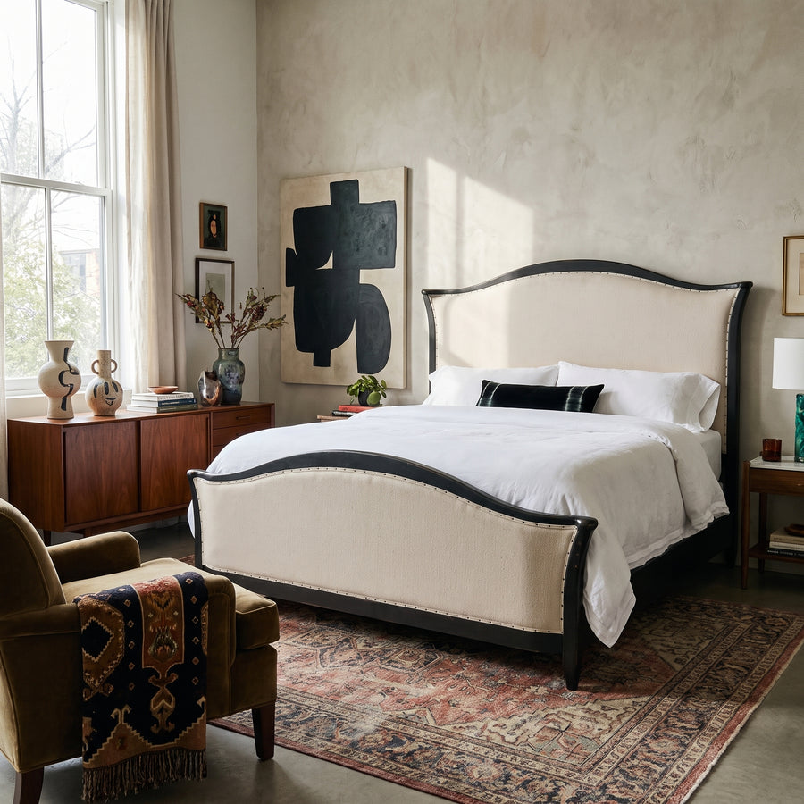 Modern bedroom with a cream upholstered bed, vintage-style rug, mid-century sideboard, and abstract wall art.