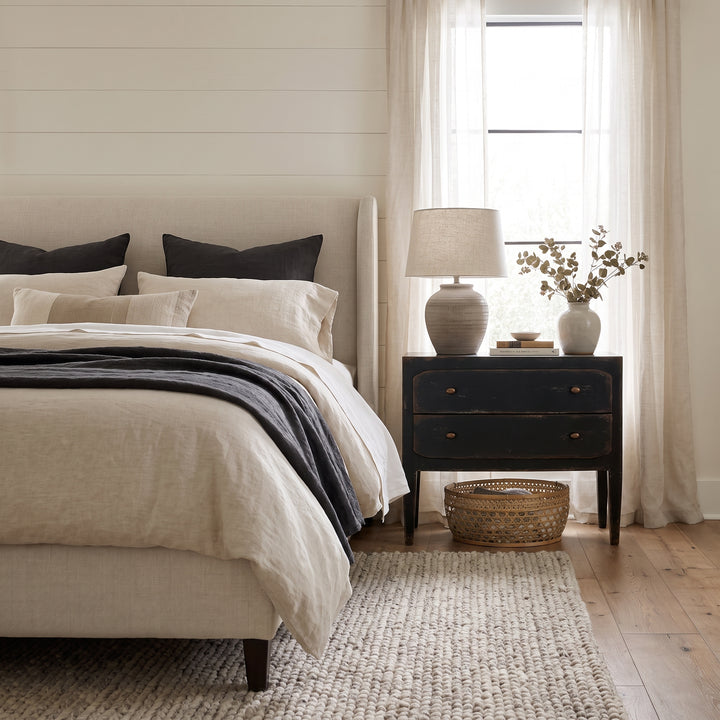 Modern farmhouse bedroom featuring a neutral linen bed, dark wood nightstand, and soft textures in a light-filled space.