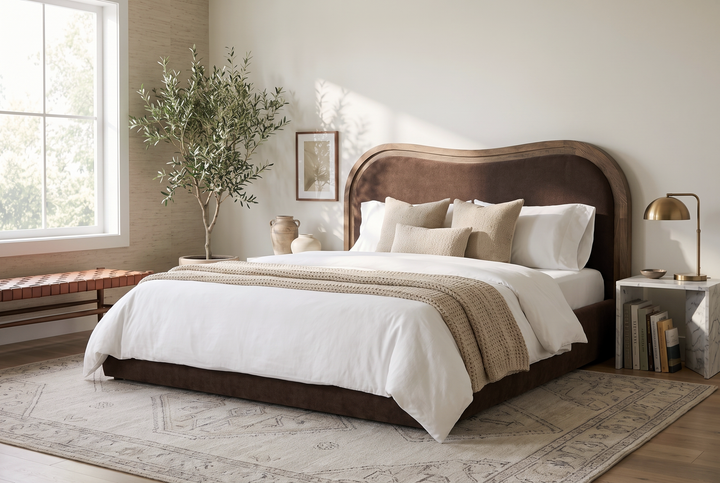 Modern bedroom with a brown upholstered arched headboard, neutral bedding, olive tree, and marble side table.